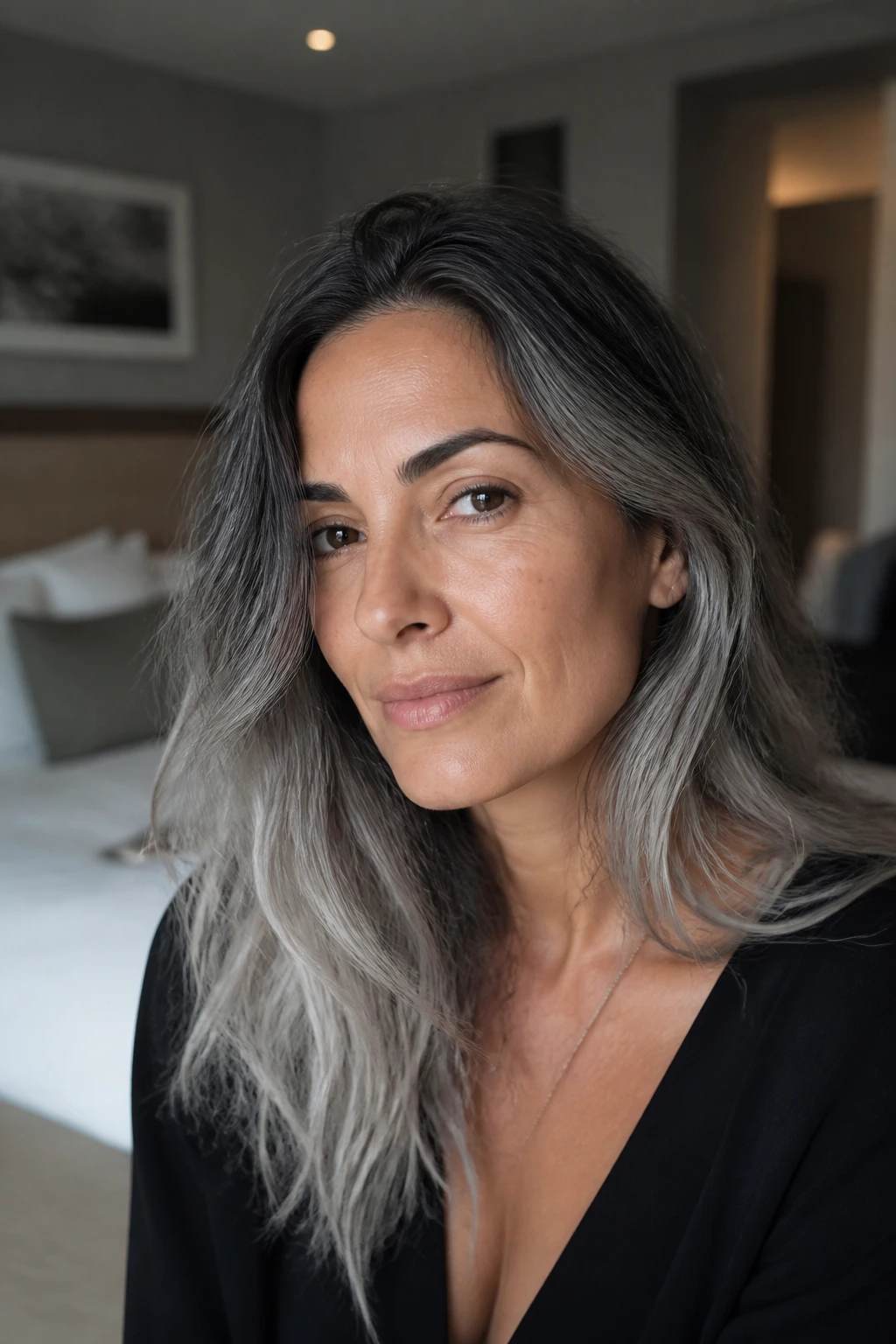 Close-up of dark rooted hair fading to cloudy silver ends on olive skin in a muted bedroom.