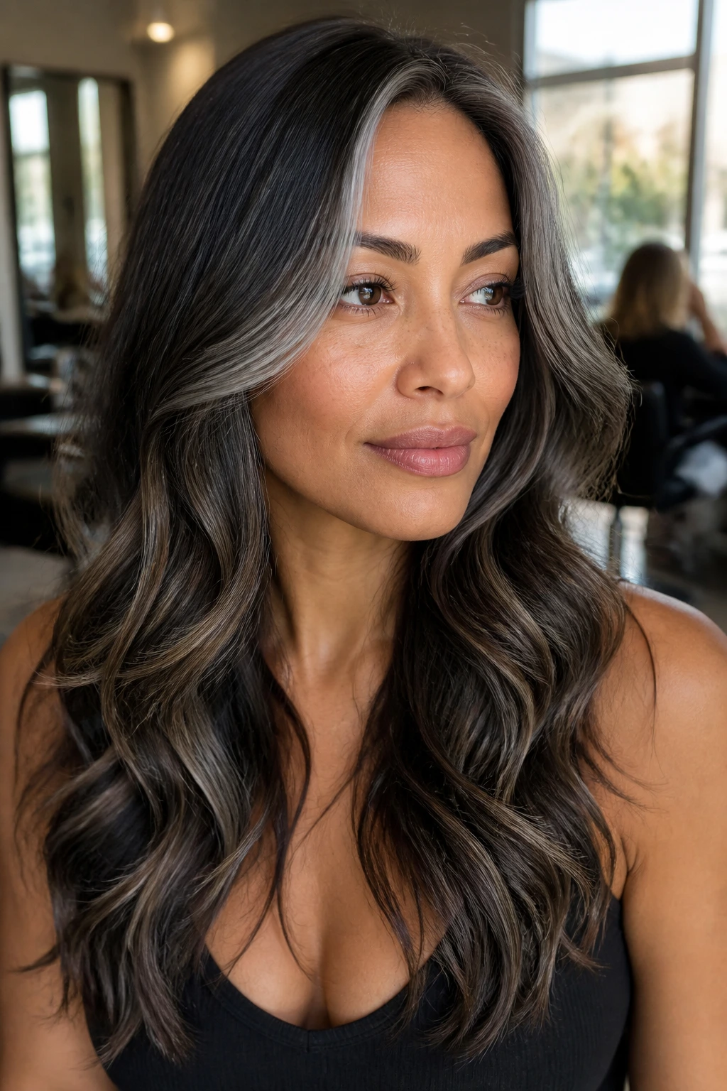 Close-up portrait of a real woman with tan skin and dark brown hair showing grey lowlights in a warm salon.